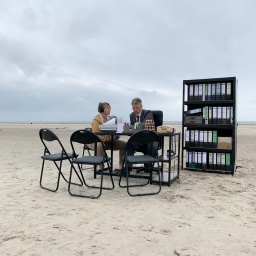 Büromöbel stehen am Strand, eine Frau und ein Mann sitzen am Bürotisch