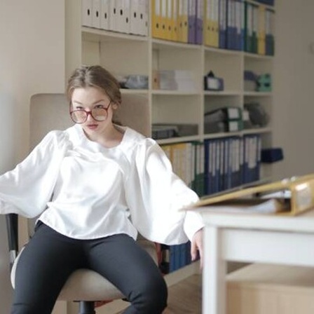 Frau sitzt mit heruntergerutschten Brille sichtlich erschöpft auf Schreibtischstuhl.