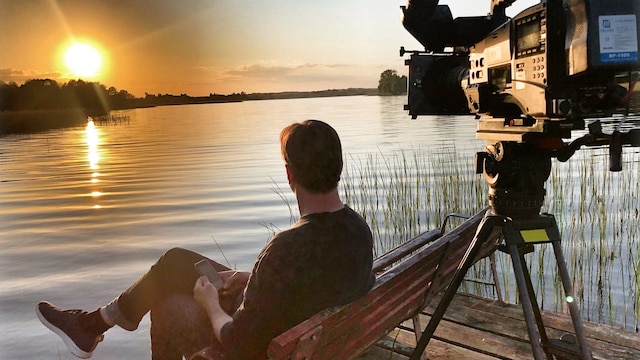 Ein Mann betrachtet am See einen Sonnenuntergang. Hinter ihm steht eine Kamera.