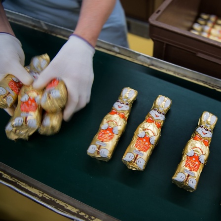Schokoosterhasen in goldener Folie liegen auf einem Fließband und werden von einer Mitarbeiterin aufgenommen. 