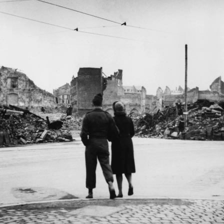 Ein US-Soldat geht mit einer deutscher Frau im Arm eine im Zweiten Weltkrieg zerstörte Straße entlang.