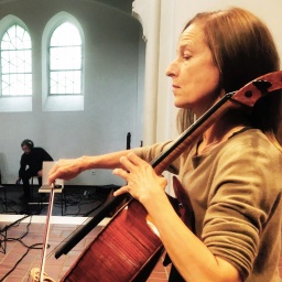 Eine Frau mittleren Alters mit langen roten Haaren spielt Cello. Im Hintergrund sieht man zwei Kirchenfenster und einen Techniker. Ein Mikrofon ist auf sie ausgerichtet.