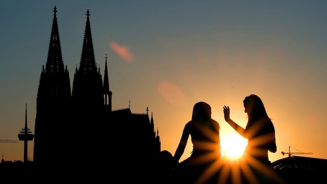 Zwei Frauen sitzen im Sonnenuntergang am Rheinboulevard vor dem Dom, man erkennt nur Umrisse