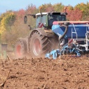 Trockener Acker mit großem Traktor samt angehängtes Gerät. Im Hintergrund ein paar gefärbte Laubbäume- Aufnahme aus Bodennähe.