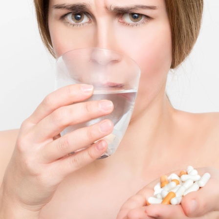 Eine grimmig aussehende Frau trinkt aus einem Wasserglas und hält Tabletten in ihren Händen