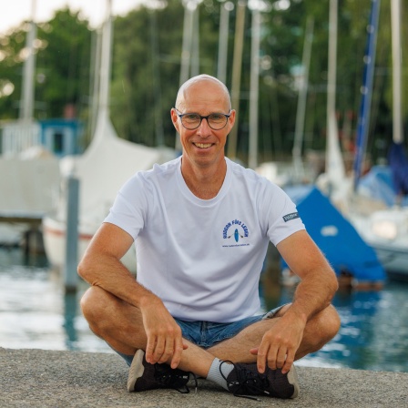 Atlantik-Soloruderer Martin Stengele sitzt im Schneidersitz, im Hintergrund sind Boote sichtbar. Martin Stengele versuchte alleine quer über den Atlantik zu rudern.