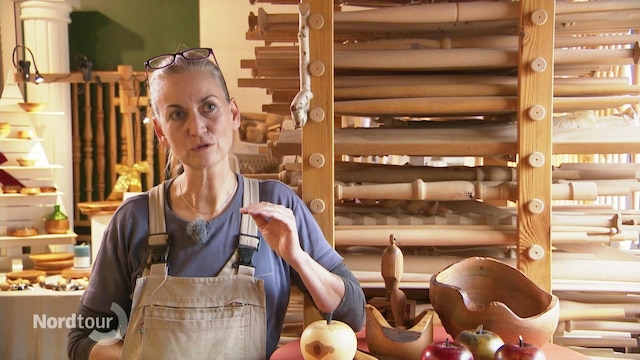 Eine Frau in einer grauen Latzhose steht in einer Holzwerkstatt.