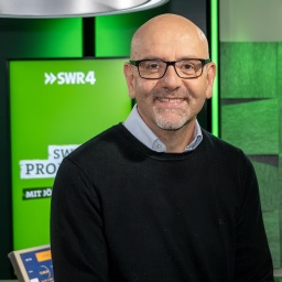 Jörg Assenheimer sitzt auf dem Tisch des Studios, in dem Assenheimers Promitalk aufgezeichnet wird, und lächelt in die Kamera. Im Hintergrund ein Monitor mit Sendungsschriftzug.