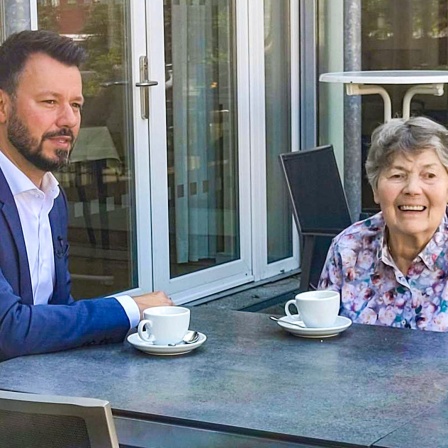 Heimleiter André Drummond und Bewohnerin Irmgard Eckert sitzen vor dem Gast- und Krankenhaus in Hamburg-Poppenbüttel an einem Tisch.