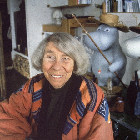 Eine ältere Frau mit grauen Haaren sitzt in einem Atelier. Es ist die Schriftstellerin Tove Jansson. Hinter ihr sind Figuren der Mumins, die Jansson erfunden hat.