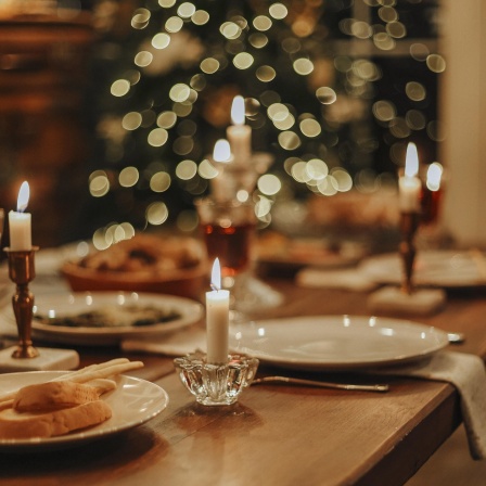 Weihnachtlich gedeckter Esstisch, mit Kerzen und Rotwein. Verschwommen im Hintergrund ein leuchtender Weihnachtsbaum.