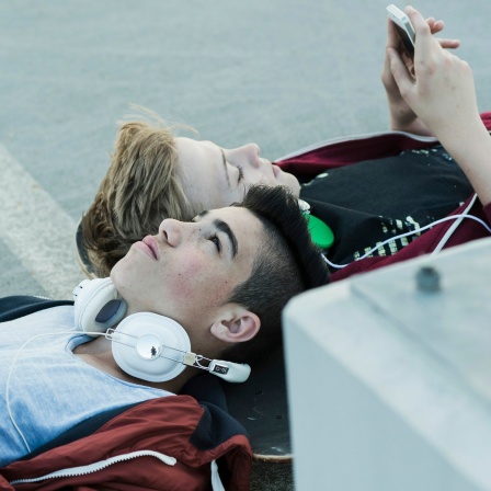 Zwei Jungs liegen mit ihren Köpfen auf einem Skateboard, sie haben ein Smartphone und Kopfhörer bei sich.