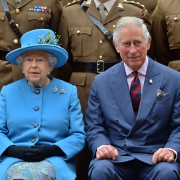 Königin Elizabeth II sitzt neben Prinz Charles. Beide sind blau gekleidet, er lächelt leicht. Hinter ihnen Menschen in Militäruniformen.