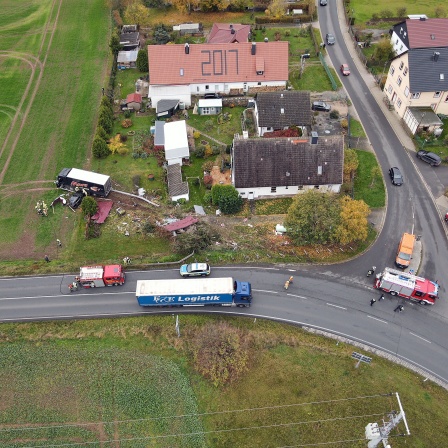 Ein Lkw steht nach einem Unfall auf einem Feld. In einem Graten hinter ihm ist eine Schneise der Verwüstung. Neben dem Lkw steht ein Auto. Es ist nach einem Zusammenprall zerstört. Auf dem Dach des Autos liegt ein Wellblech.