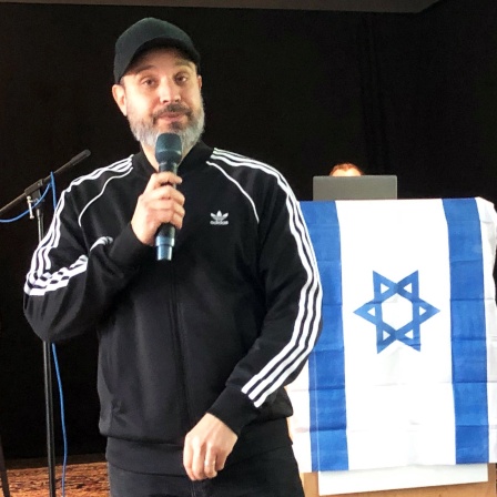 Ben Salomo, in Israel geborener Rapper und Referent zum Thema Antisemitismus, spricht vor einer israelischen Flagge bei einem Vortrag vor Schülerinnen und Schülern.