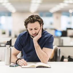 Ein Mann stützt sich auf seinen Ellenbogen und betrachtet ein aufgeschlagenes Buch auf einem Schreibtisch