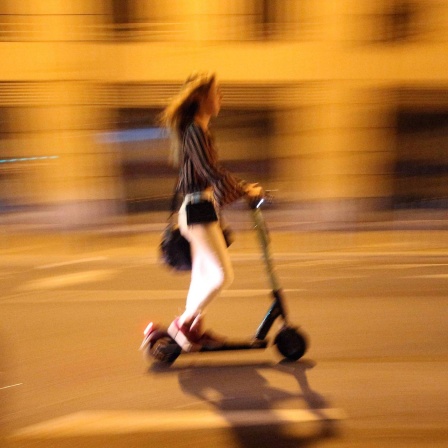 Eine junge Frau fährt abends auf einem E-Scooter durch die Stadt, der Hintergrund ist verschwommen.