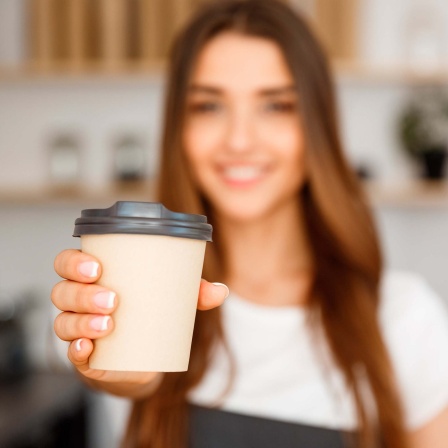 Ein junger Mensch mit Schürze reicht einen Kaffebecher in Richtung Kamera