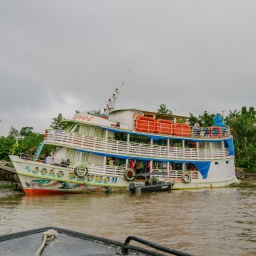 Ein zweitstöckiges Schiff liegt am Ufer eines großen tropischen Flusses, über einen Steg gehen Menschen an Bord