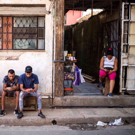 Eine Straßenszene in Kubas Hauptstadt Havana: Fünf Personen, drei Männer, ein Mädchen und eine Frau, sitzen an einem Gehweg an einer Straße und schauen auf ihre Smartphones.