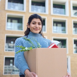 Eine junge Frau steht lächelnd mit einem Umzugskarton in den Händen vor einem Studentenwohnheim.