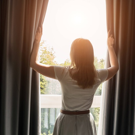 Frau öffnet Vorhänge in einem Zimmer am sonnigen Morgen.
