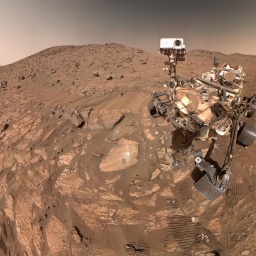 Ein Foto zeigt eine steinige rote Landschaft auf dem Mars, rechts sieht man den Forschungsrover Perseverance und Abrücke von dessen Rädern im Sand.