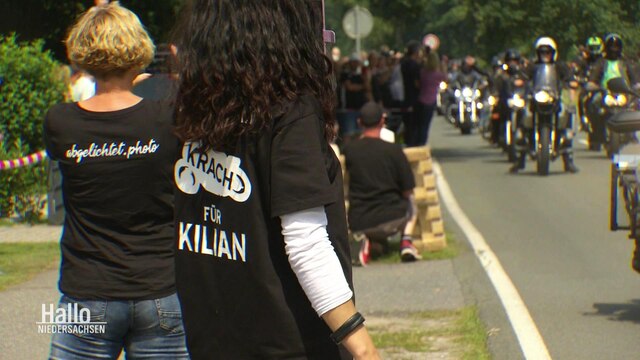 Menschen stehen am Straßenrand, während Motorradfahrer an ihnen vorbei fahren. Die Menschen an der Seite tragen T-Shirts auf deren Rücken der Name "Kilian" geschrieben steht.