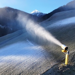 Das Bild zeigt einen Bergabhang ohne echten Schnee. Eine gelbe Schneekanone schießt weißen Kunstschnee auf die abschüssige Wiese.  