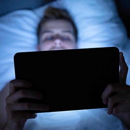 Foto eines Mannes, der auf dem Rücken im Bett liegt und auf einem Tablet einen Film schaut.