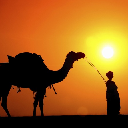 Mann mit Kamel und Frauen, die Wassertöpfe tragen, bei Sonnenuntergang durch Sanddünen.