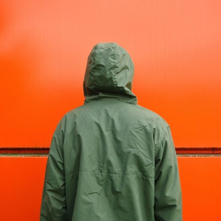 Ein Mann in einer grünen Regenjacke steht mit dem Gesicht zu einer roten Wand