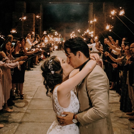 Ein Hochzeitspaar küsst sich, während die Gäste dahinter Spalier stehen und brennende Wunderkerzen halten. 