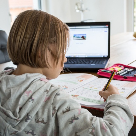Ein junges Mädchen sitzt am Wohnzimmertisch vor einem Laptop und schreibt in ein Schulheft.