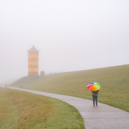 Eine Frau geht mit Regenschirm vor einem Leuchtturm in Pilsum spazieren.