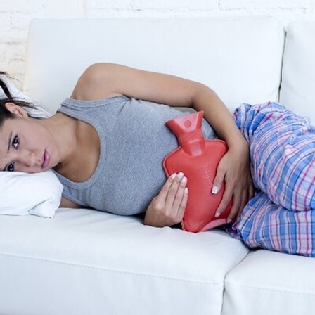 Eine Frau liegt mit einer Wärmflasche vor dem Bauch auf einem weißen Sofa.