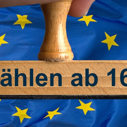 Ein symbolischer Holzstempel mit der Aufschrift "Wählen ab 16", wird von einer Hand im Anschnitt vor einer EU-Flagge gehalten.