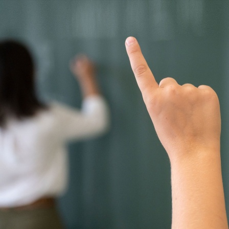 Finger eines Kindes zeigt auf, im Hintergrund steht eine Lehrerin vor der Tafel