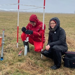 Eine Frau und ein Mann hocken auf einem Feld vor Gerätschaften.