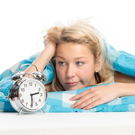 Junge Frau liegt schlaflos im Bett und starrt auf den Wecker: Es ist wichtig, dass Insomnien, also Ein- und Durschschlafprobleme, behandelt werden, weil sie andere Erkrankungen nach sich ziehen können, etwa Herz-Kreislauf-Erkrankungen und Depressionen