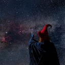 Eine Person mit blauem Umhang, rotem Spitzhut und fingerlosen Handschuhen steht vor einem nächtlichen Sternenhimmel.