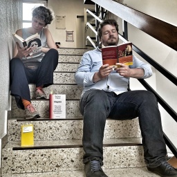 Lisa Kreißler und Alexander Solloch sitzen lesend auf Stufen eines Treppenhauses. 