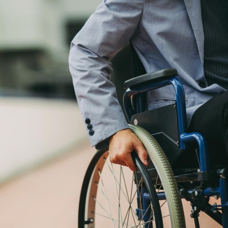 Ein Mann mit Sakko sitzt in einem Rollstuhl vor einem Geschäftsgebäude. Zu sehen ist nur seine Hand an einem Rad des Rollstuhls