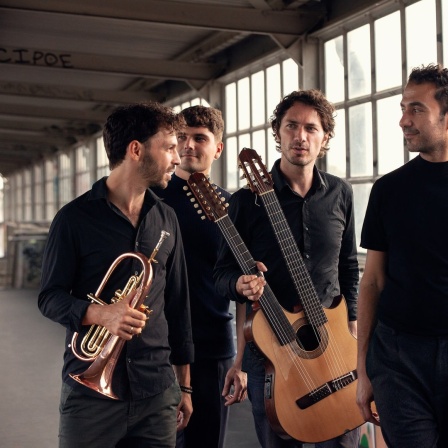 Eine Gruppe von vier Männern mit Musikinstrumenten posiert in einer Halle vor der Kamera.