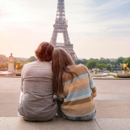 Ein Mann und eine Frau reiferen Alters sitzen eng aneinandergeschmiegt und betrachten den Eiffelturm in Paris. Sie sind in Rückenansicht zu sehen.