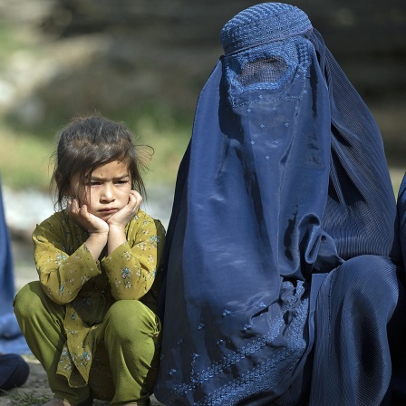 Ein Mädchen in Afghanistan hockt zwischen zwei verhüllten Frauen auf sandigem Boden.