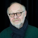 Der Regisseur Jürgen Flimm, mit Brille und weißem Bart, steht vor einem dunklen Hintergrund und schaut freundlich konzentriert in die Kamera.