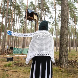 Eine Klimaaktivistin steht mit dem Rücken zur Kamera und schaut auf Baumhäuser als Teil des Protestcamps in einem Kiefernwald bei Seehausen. Auf einem Transparant steht A14 - Schnee von gestern.