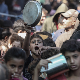 Dieses Foto zeigt eine Menschenmenge, in der viele Personen, vor allem Kinder, sichtbar hungrig und verzweifelt wirken. Im Vordergrund hebt ein Junge mit ernstem Gesichtsausdruck eine Metallplatte hoch, möglicherweise um auf sich aufmerksam zu machen oder um Nahrung zu erbitten.