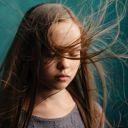 Trauernden Kindern und Jugendlichen beizustehen führt oft auch Erwachsene an emotionale Grenzen. Zu sehen: Ein Mädchen mit geschlossenen Augen und wehenden Haaren. Der Hintergrund ist eine grüne Wand. 